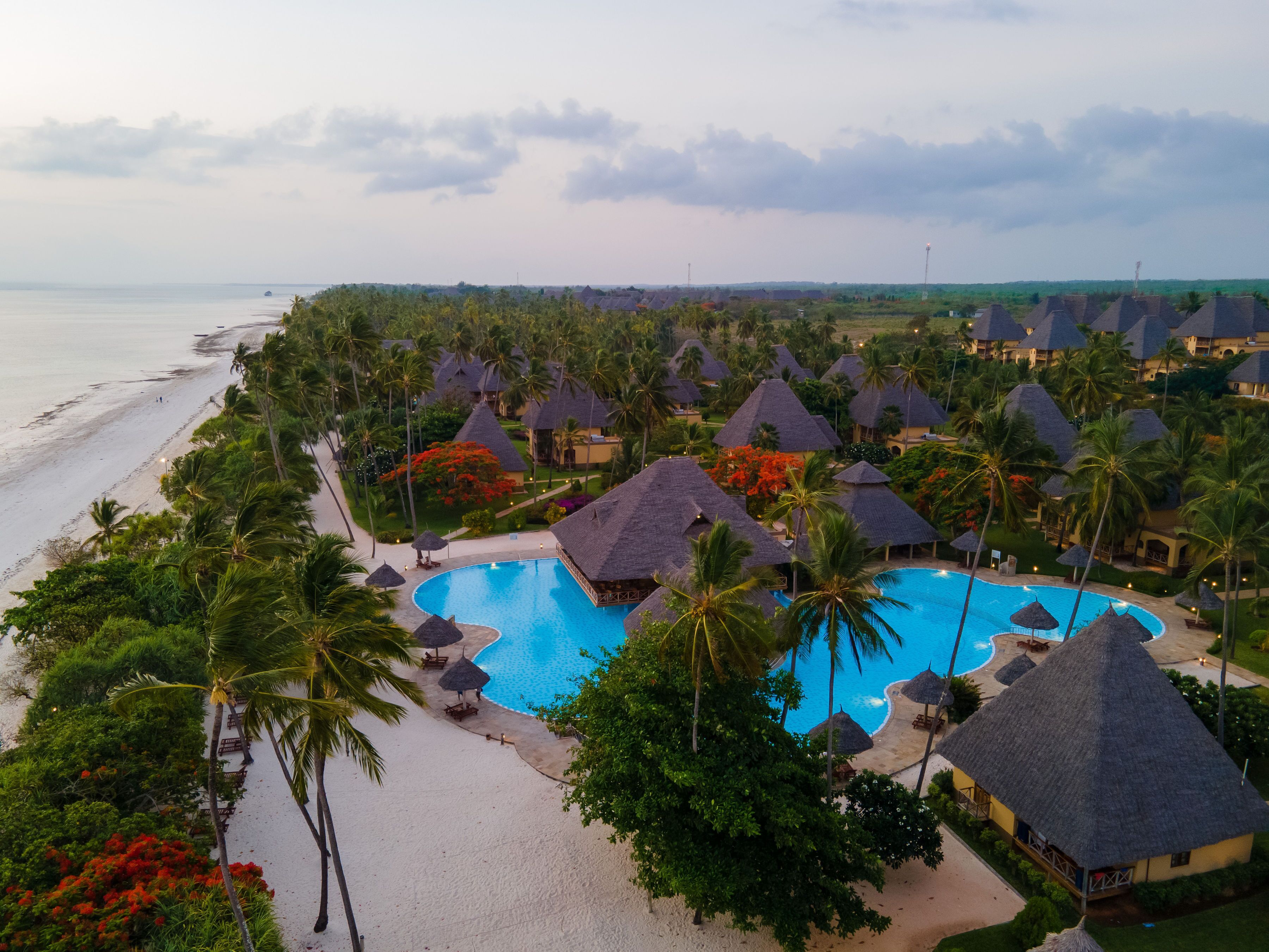 A resort with a central swimming pool, beach, and thatched-roof bungalows.