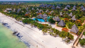 Vista aérea de un resort con varias cabañas, una piscina central y una playa con un velero.