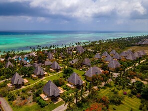 Vue aérienne d’un complexe hôtelier avec des bungalows aux toits de chaume, une piscine et une vue sur l’océan et ses eaux d’un bleu limpide.