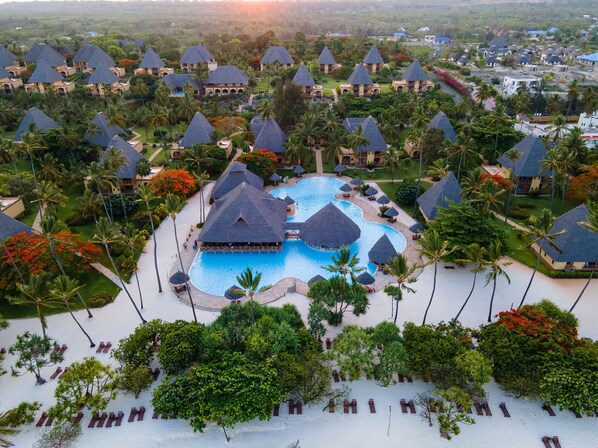 Vue aérienne d’un complexe hôtelier comprenant plusieurs bâtiments de style bungalow, une piscine centrale et des palmiers environnants.