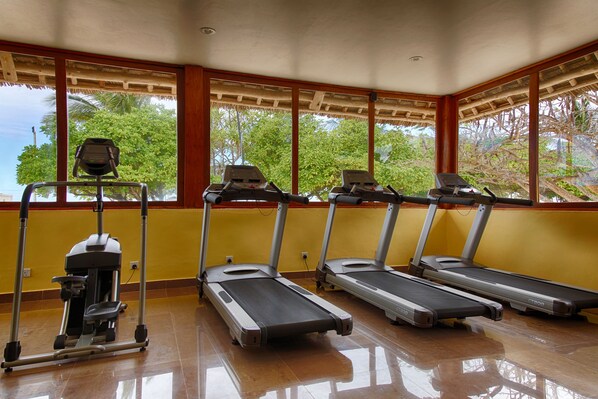 A gym with treadmills and a stationary bike, large windows with a view of trees, and yellow walls. - Neptune Pwani Beach Resort & Spa Zanzibar (Kiwengwa)