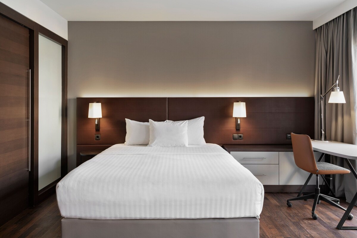 A hotel room with a large bed, two bedside lamps, a desk with a chair, and a wardrobe.