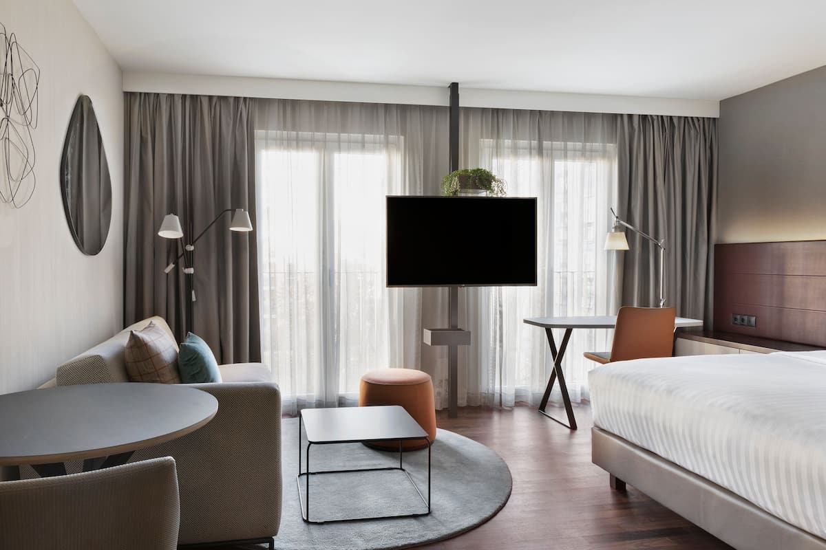A modern hotel room with a bed, a round dining table, a sofa, a TV, and a small table with lamps.