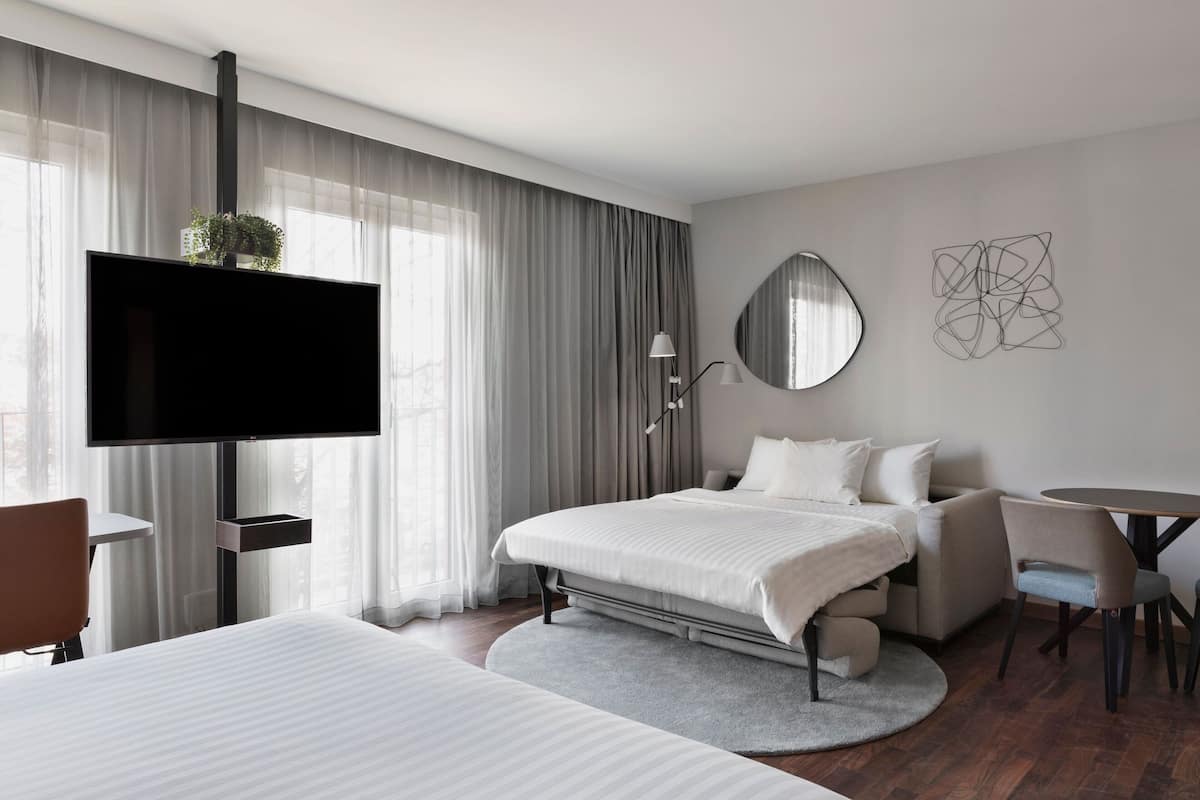 A modern hotel room with a bed, a sofa, a TV, a desk, and a dining area.