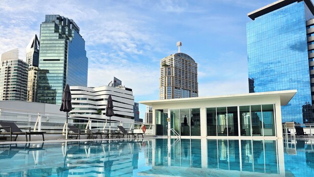 A rooftop pool with lounge chairs and a glass-enclosed cabana, surrounded by modern high-rise buildings. - The Bless Hotel and Residence (Bangkok)