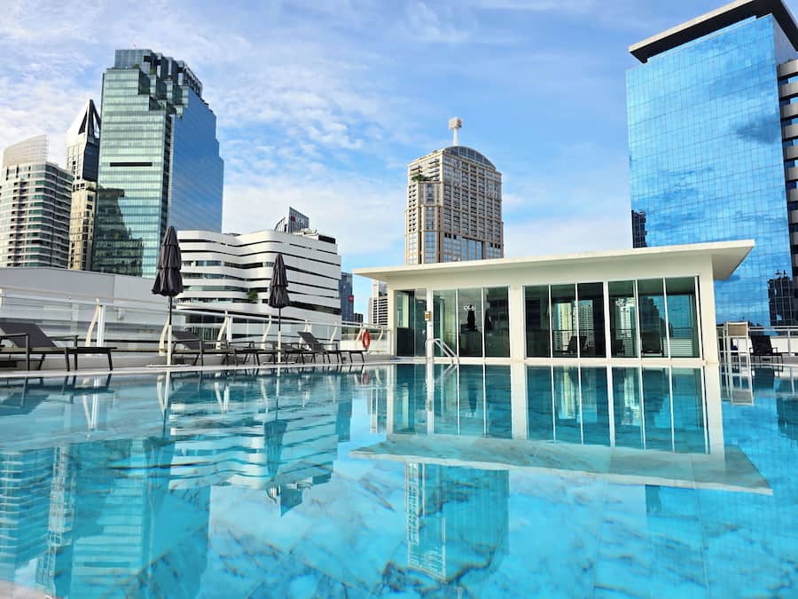 Uma piscina no terraço com espreguiçadeiras e um bangalô com paredes de vidro, cercada por edifícios modernos e altos.