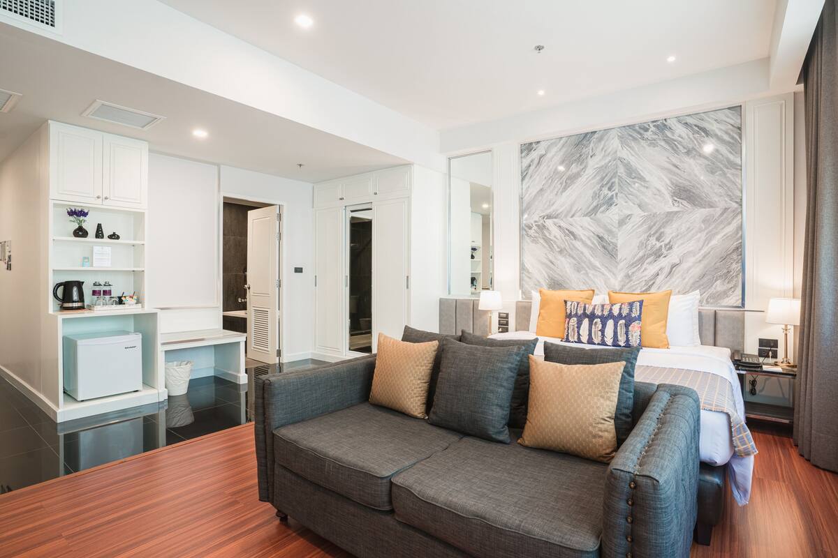 A modern hotel room with a grey sofa, a bed with multiple pillows, a marble-patterned wall, and a small kitchenette area.