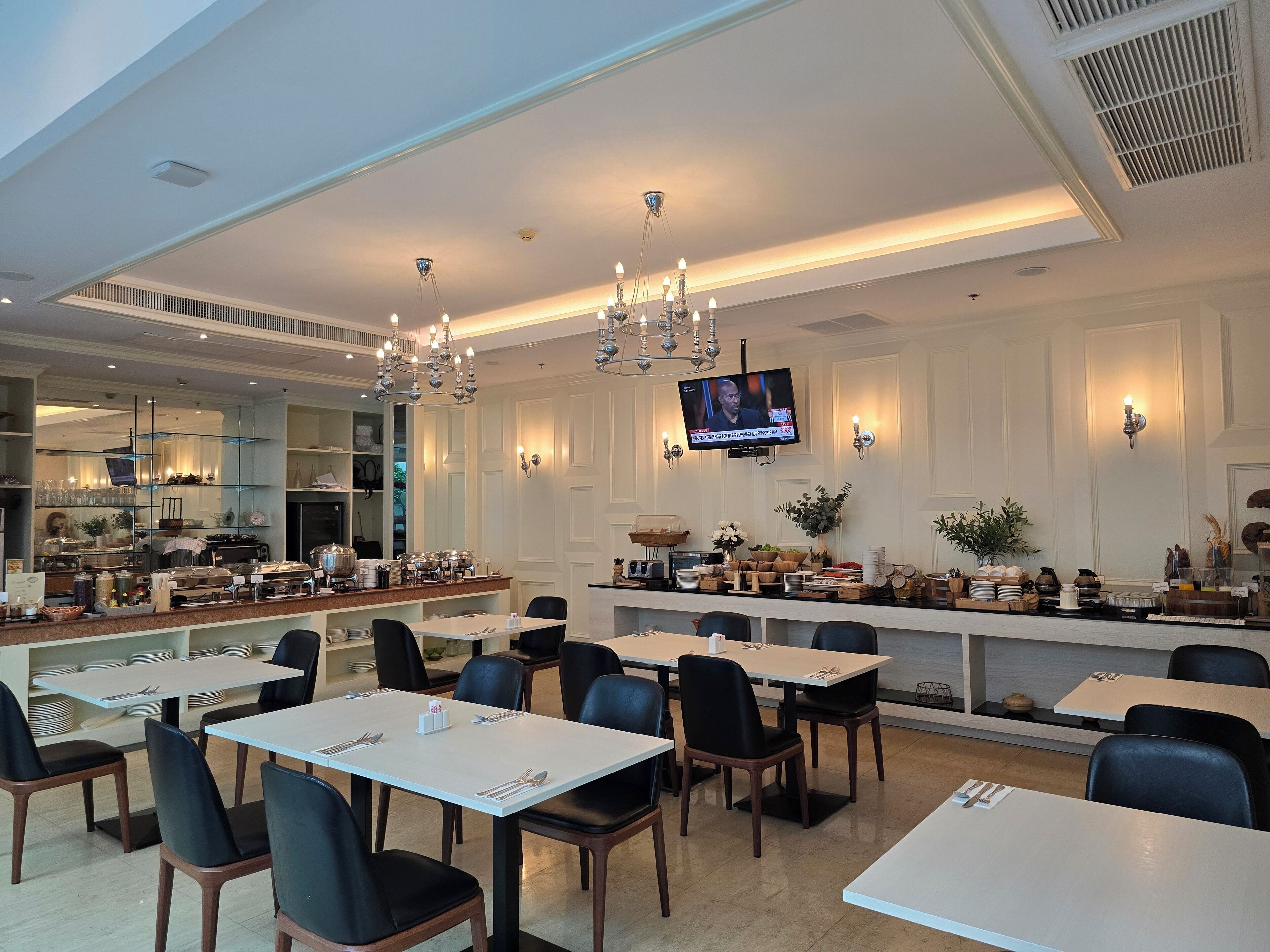 A modern dining area with multiple tables and chairs, a television mounted on the wall, and a buffet counter with various food items.
