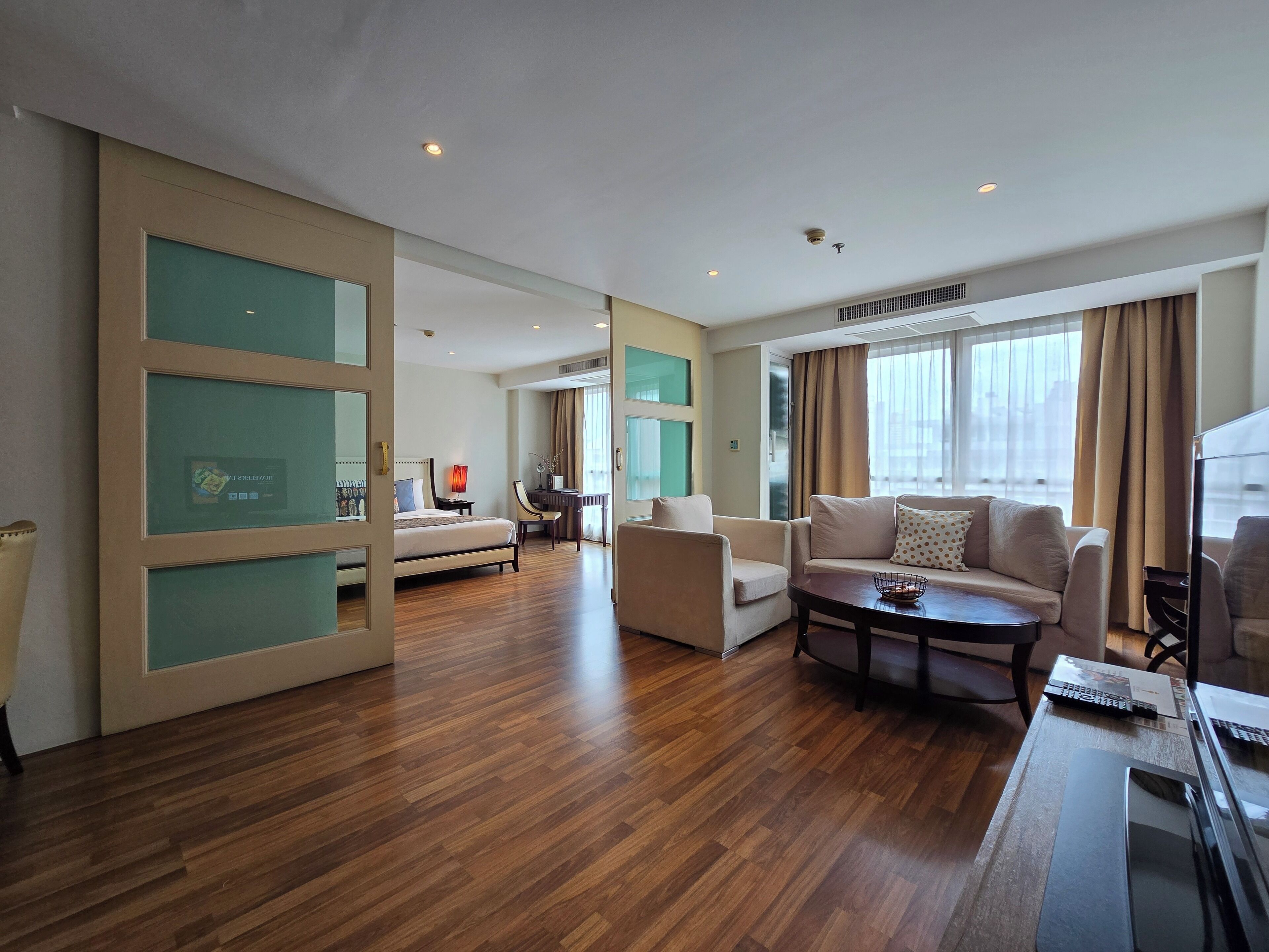 A spacious hotel room with a large bed, a sitting area with a sofa and armchairs, a desk with a computer, and a large window with curtains.