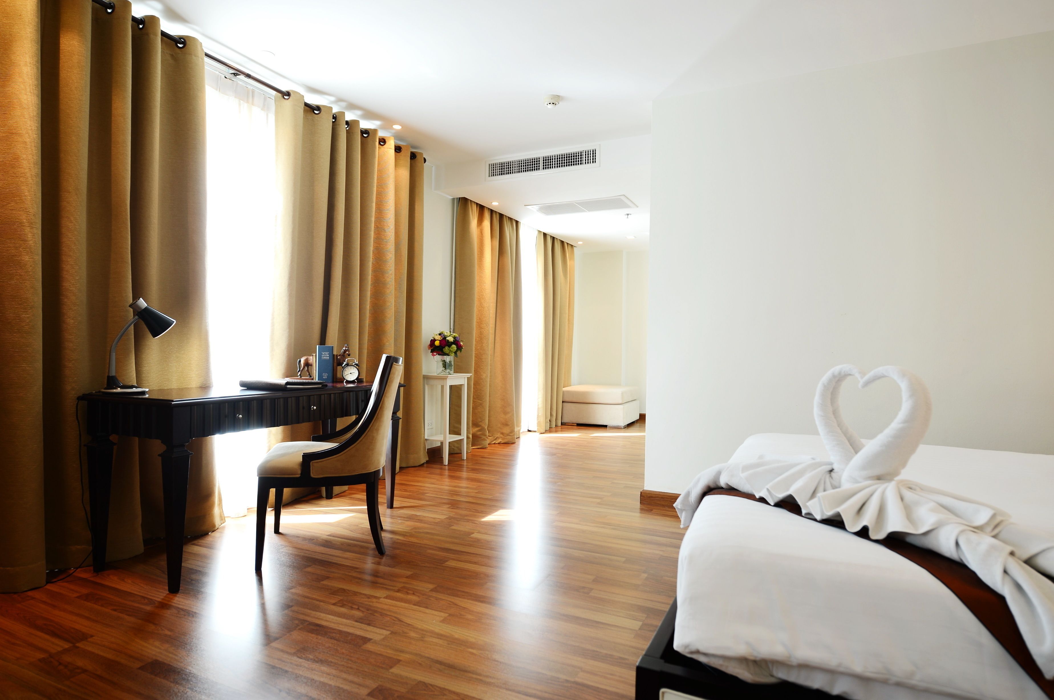 A hotel room with a wooden floor, a desk with a lamp, a chair, a bed with a heart-shaped towel, and a window with curtains.