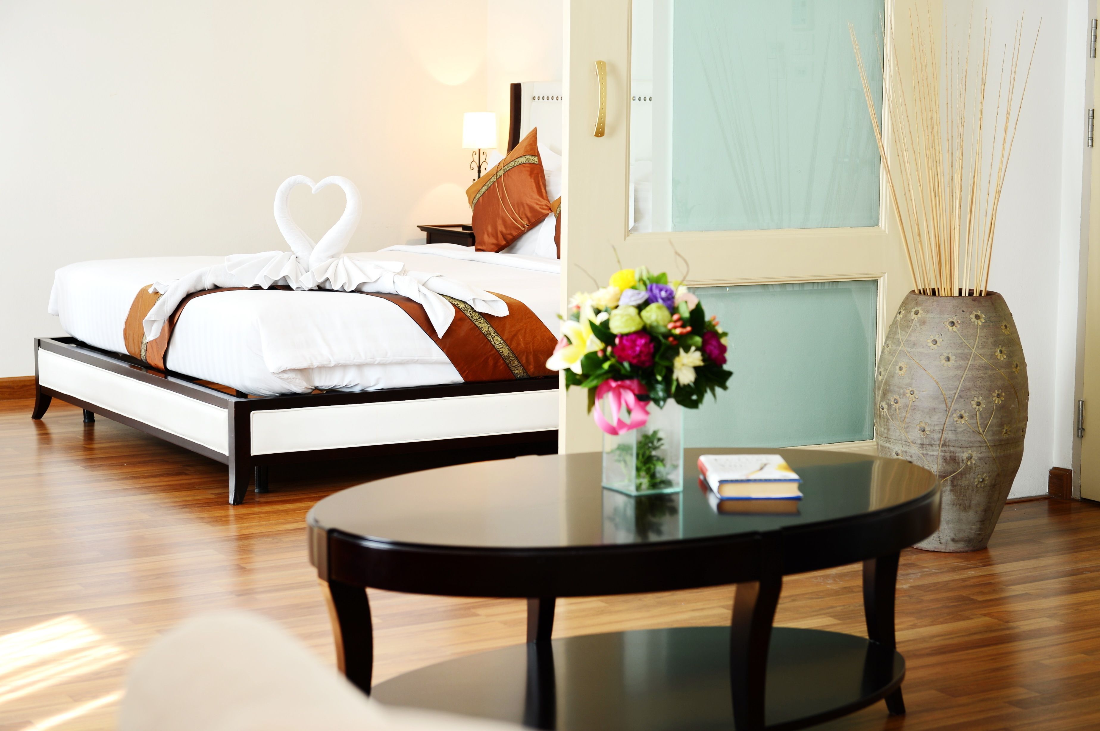 A neatly made bed with a heart-shaped towel, a bedside table with a book, a vase with flowers, and a large vase with reeds.