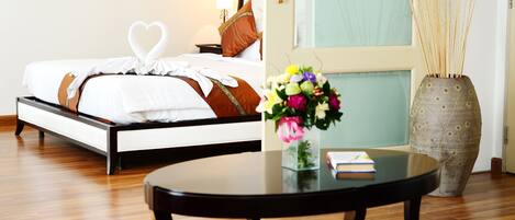 A neatly made bed with a heart-shaped towel, a bedside table with a book, a vase with flowers, and a large vase with reeds.