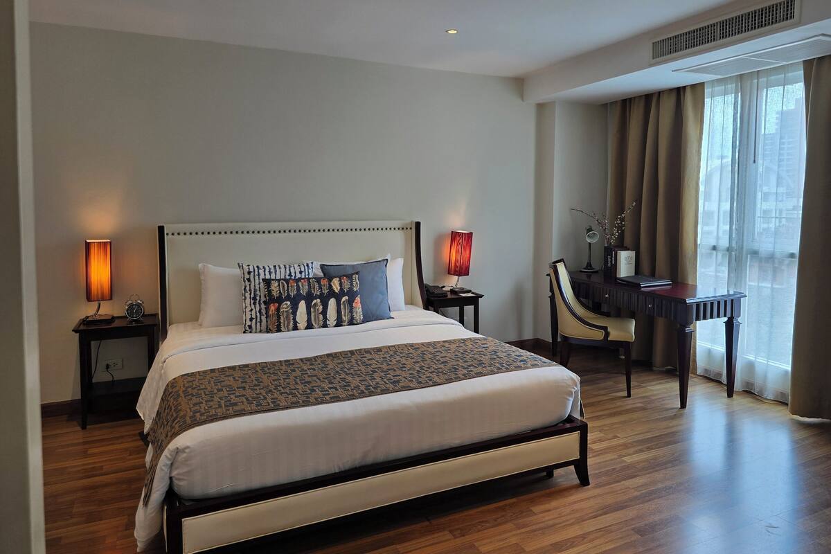 A hotel room with a large bed, bedside lamps, a desk with a chair, and a view of the city through the window.