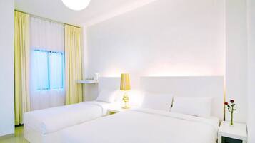 A hotel room with two beds, a bedside table with a lamp, a window with curtains, and a vase with flowers on the nightstand.