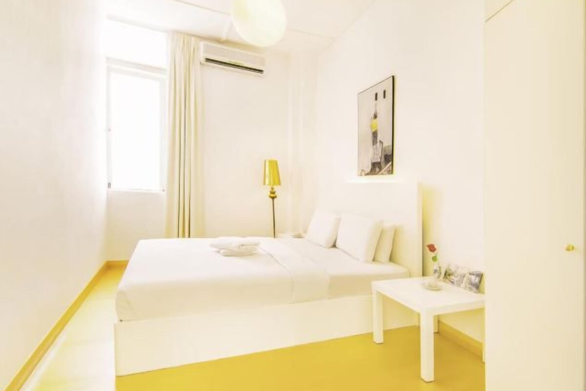 A bright, white room with a bed, bedside table, and a picture on the wall.
