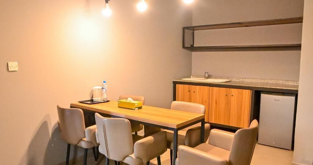 A dining area with a wooden table and six chairs, a small kitchen with a sink and cabinets, and a refrigerator.