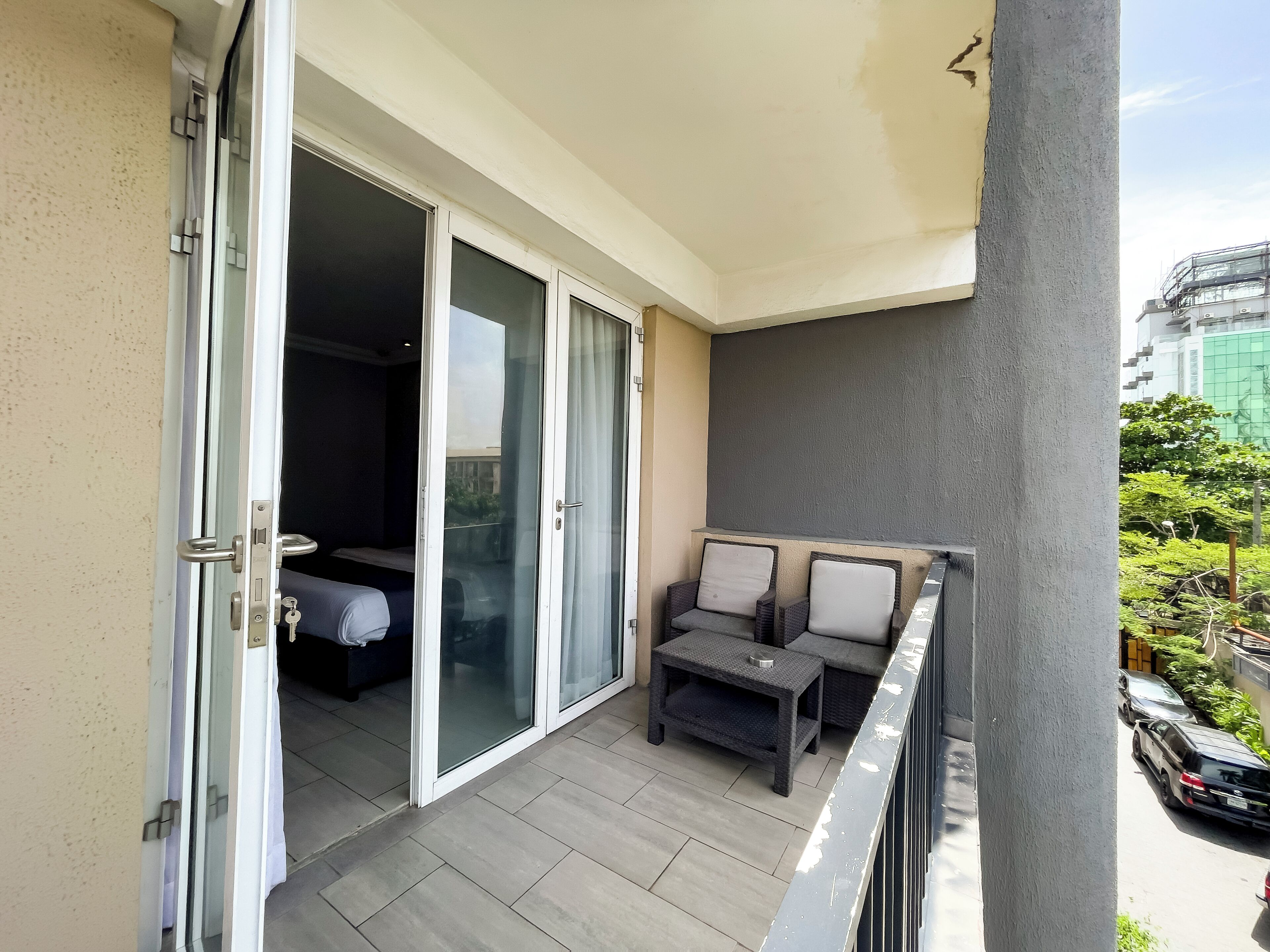 A balcony with a sliding glass door leading to a bedroom, a small outdoor seating area with two chairs and a table, and a view of a cityscape with buildings and greenery.