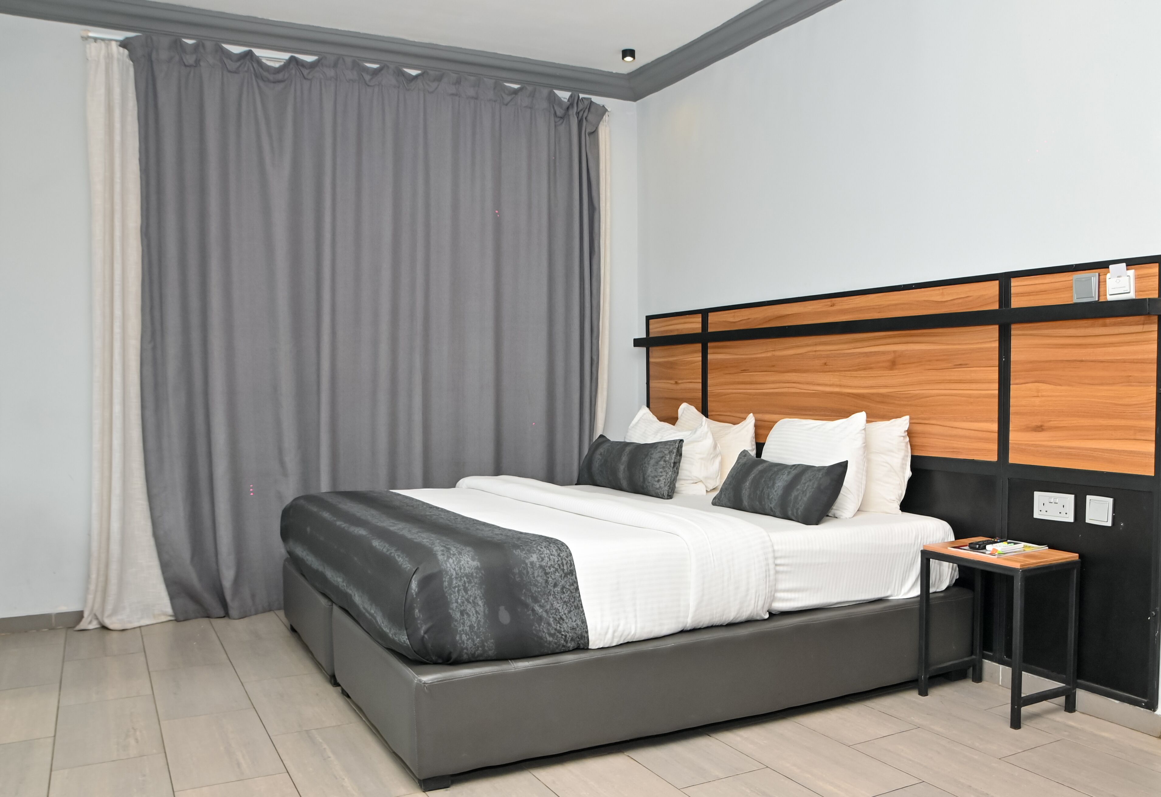 Une chambre à coucher moderne avec un grand lit, des rideaux gris, une tête de lit en bois et une petite table de chevet.