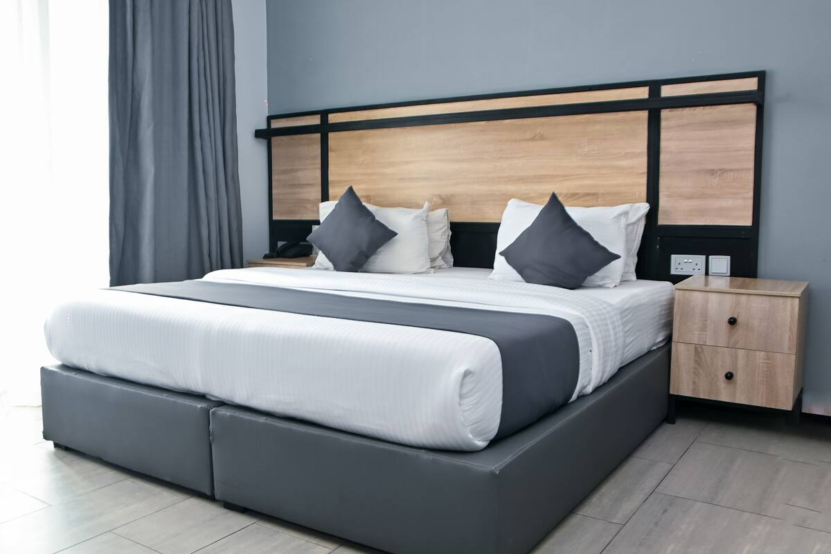 A modern hotel room with a large bed, grey and white bedding, a wooden headboard, a bedside table with a lamp, and a nightstand with a power outlet.