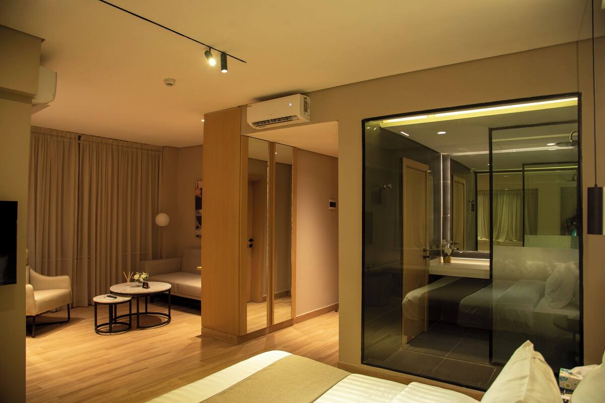 A modern hotel room with a bed, a seating area with a small table, a glass-enclosed shower, and a large mirror.