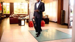 A man in a suit holding a suitcase and a phone in a hotel lobby with seating areas and a chandelier. - The Blowfish Hotel (Lagos)