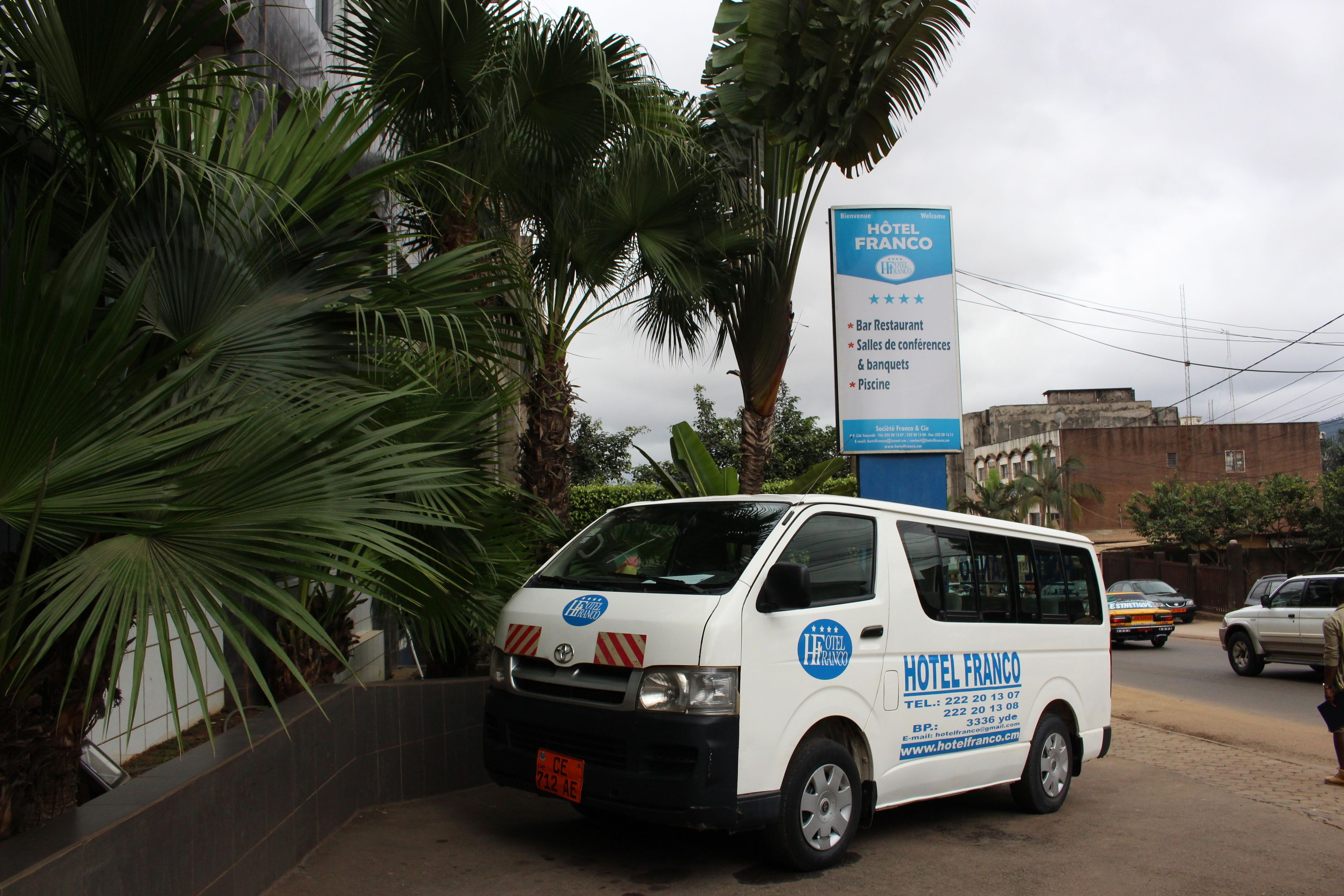 A white van with "HOTEL FRANCO" branding parked in front of a signboard listing amenities like a bar restaurant, conference halls, and a pool.