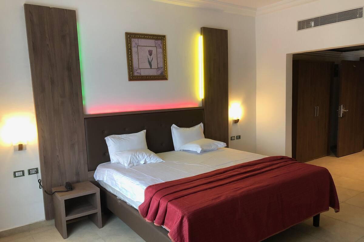 A hotel room with a large bed, a bedside table, a nightstand, a framed picture on the wall, and a door leading to another room.