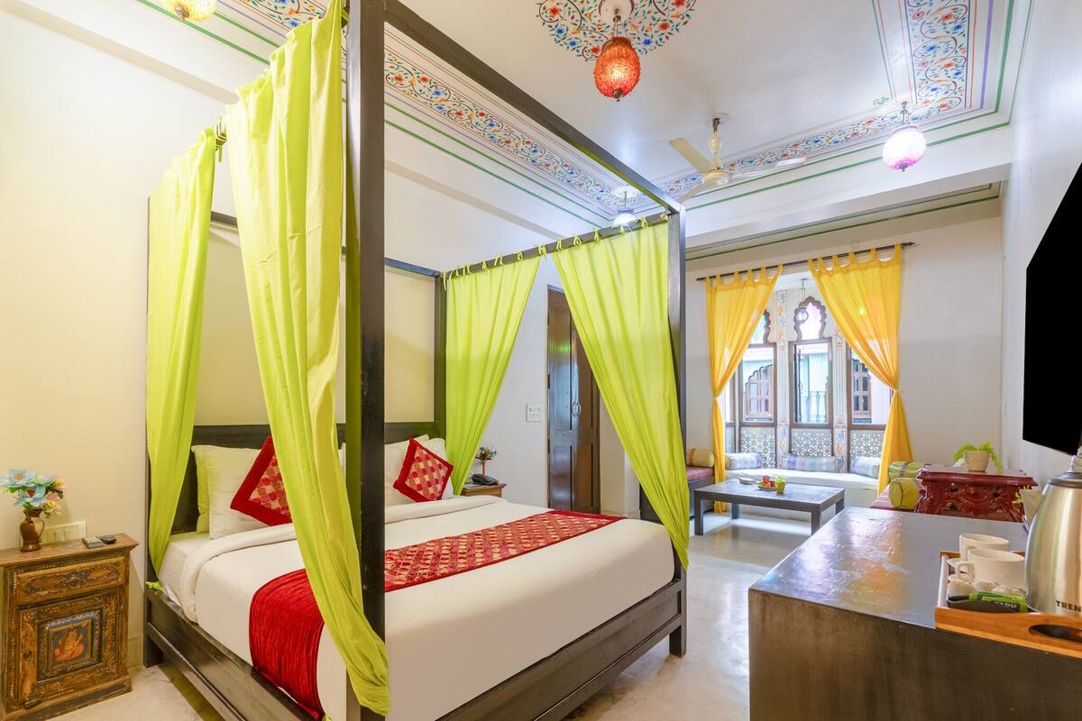 A four-poster bed with green drapes, a wooden nightstand with a statue, a bedside table with a lamp, and a view of a courtyard through a window with yellow curtains.