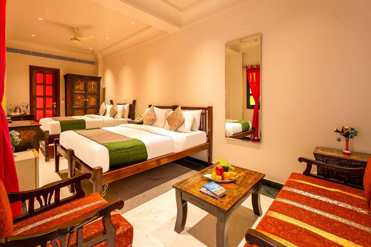 A hotel room with two beds, a wooden table with a fruit bowl, a mirror, and a red curtain.
