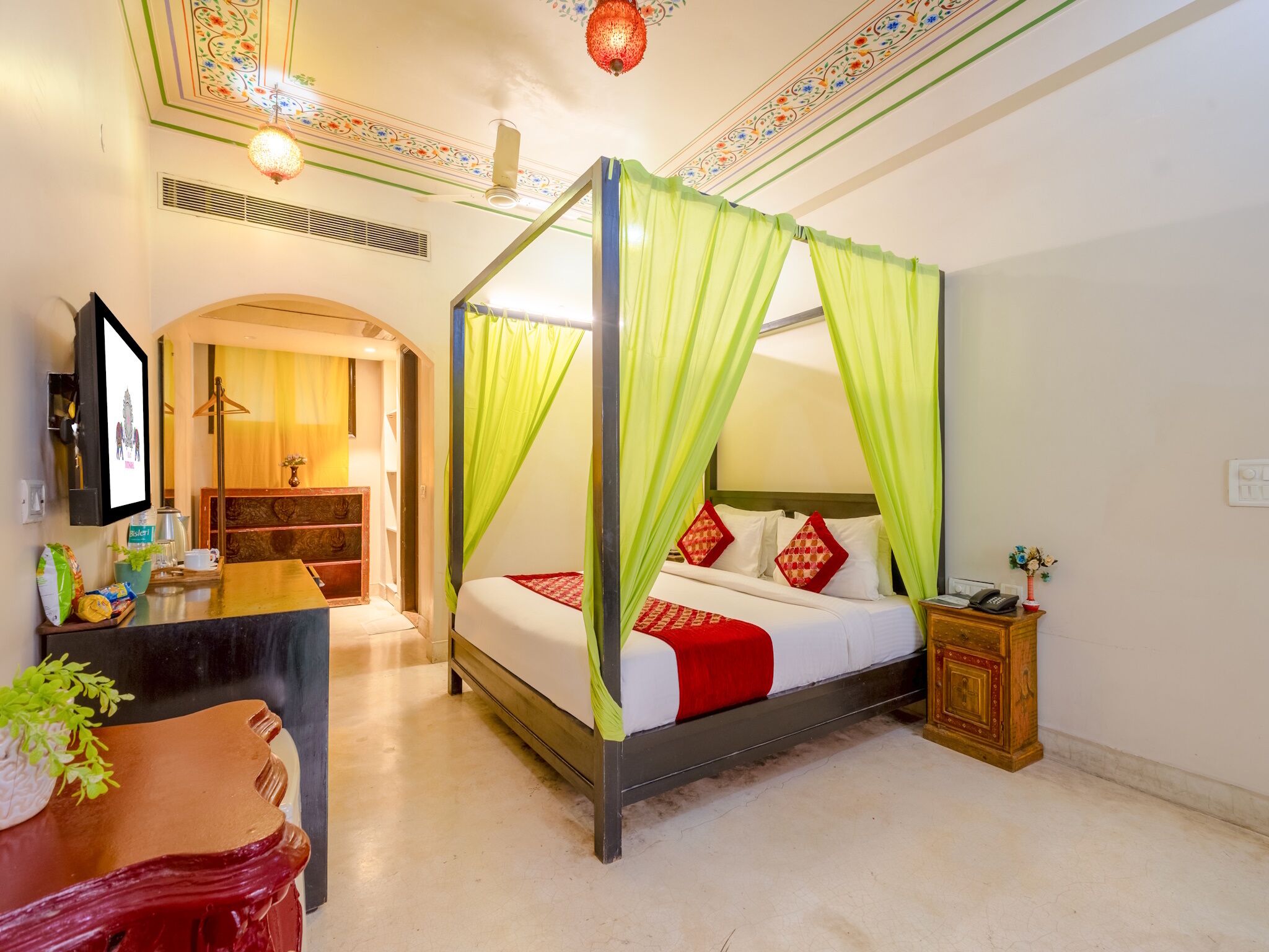 A four-poster bed with green canopy, a wooden nightstand, a desk with a chair, and a small table with a plant.