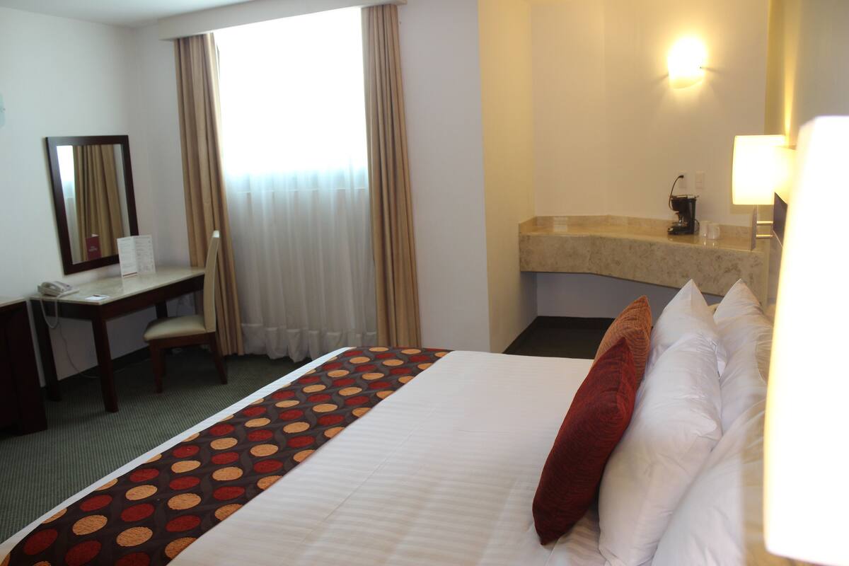 Habitación de hotel con cama, escritorio, silla, espejo, ventana con cortinas, lavamanos y lámpara.
