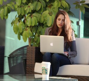 Une personne est assise sur un canapé blanc, un ordinateur portable et une tasse de Starbucks sont posés sur une table en verre.