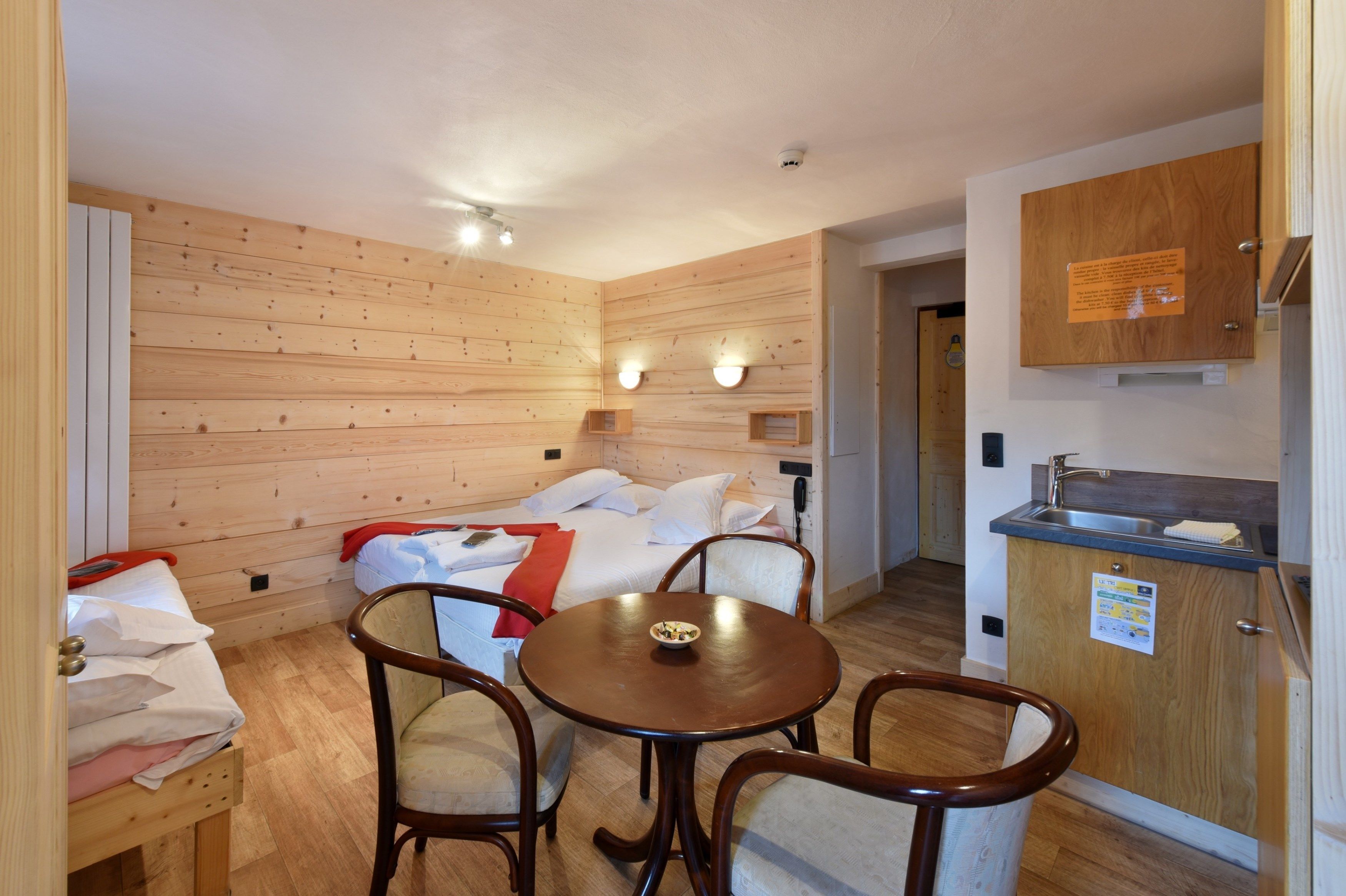 A cozy room with a bed, a small table with chairs, a wooden cabinet with a sink, and a bulletin board on the wall.