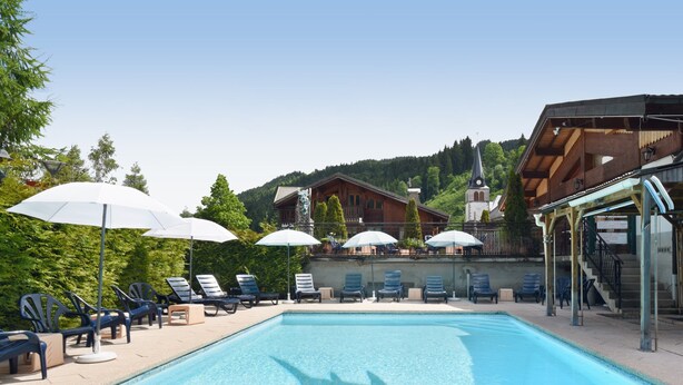 A swimming pool with lounge chairs and umbrellas, a wooden building with a staircase, and a church in the background. - Loc Hotel Alpen Sports (Les Gets)