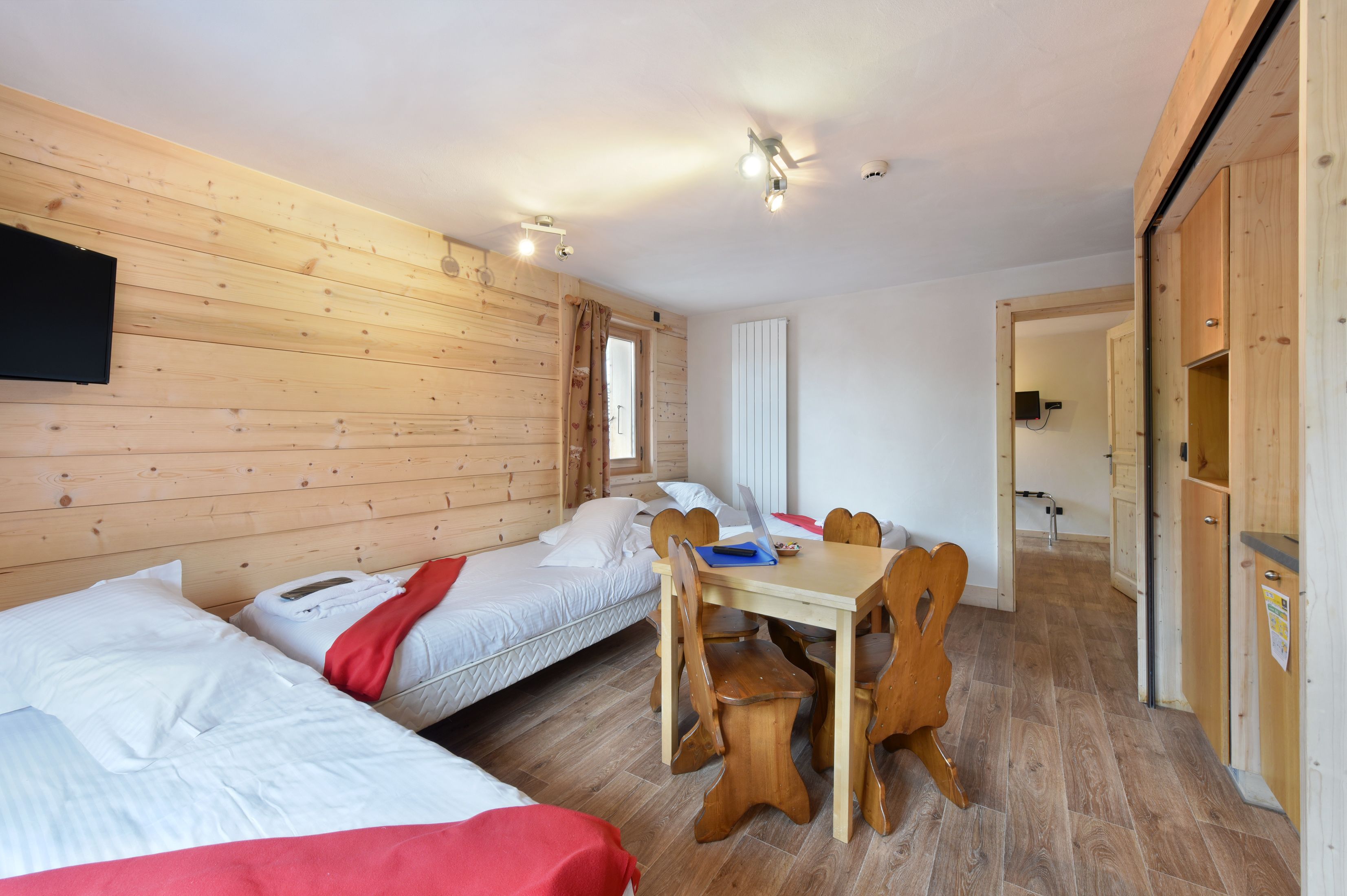 A cozy room with two beds, a wooden dining table with chairs, and a television mounted on the wall.