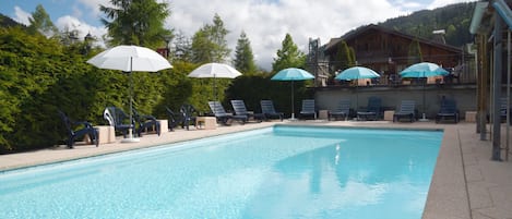 Una piscina con lettini e ombrelloni, circondata da alberi e un edificio con un balcone.