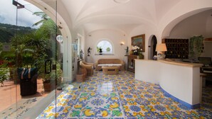 A spacious room with a tiled floor, a sofa, a wicker chair, a glass door leading outside, and a small desk with a lamp. - Semiramis Hotel De Charme (Forio)