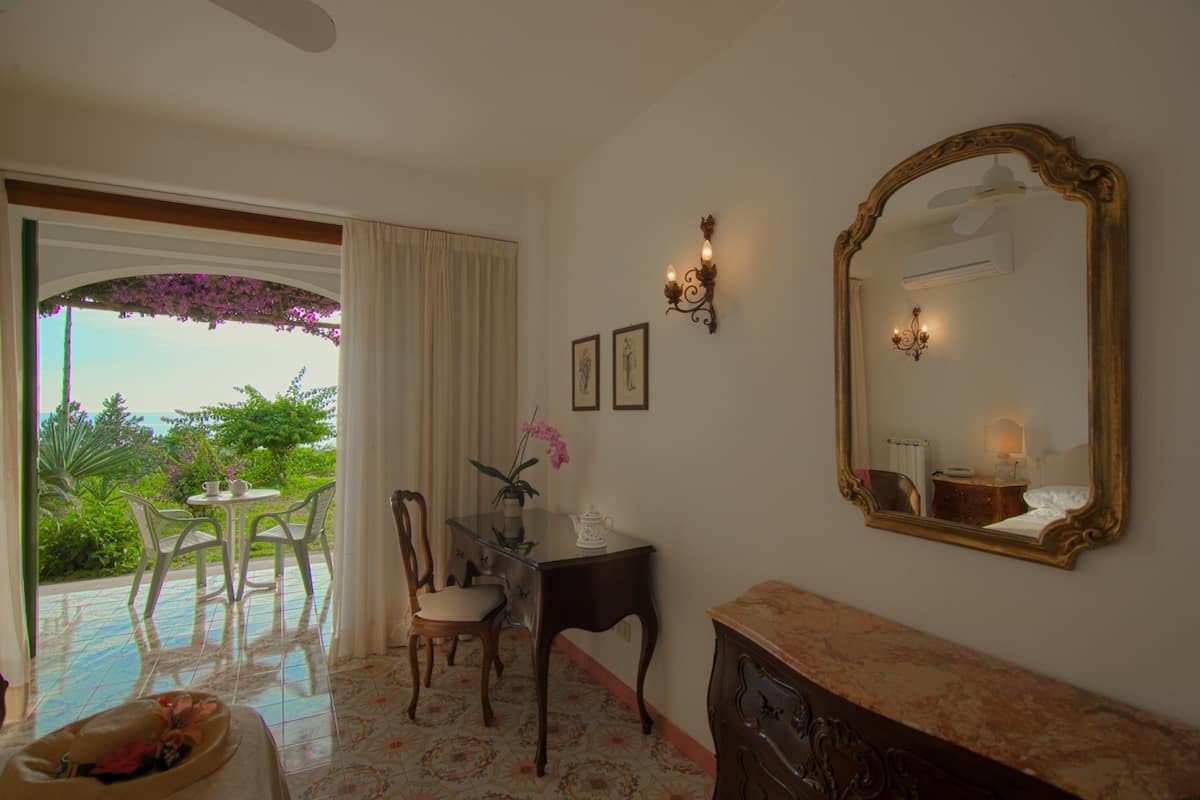 A room with a dining table, chairs, a mirror, and a view of greenery outside.