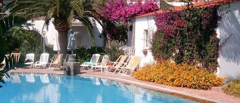 A swimming pool with lounge chairs and a statue, surrounded by flowering plants and a white building with a red roof.
