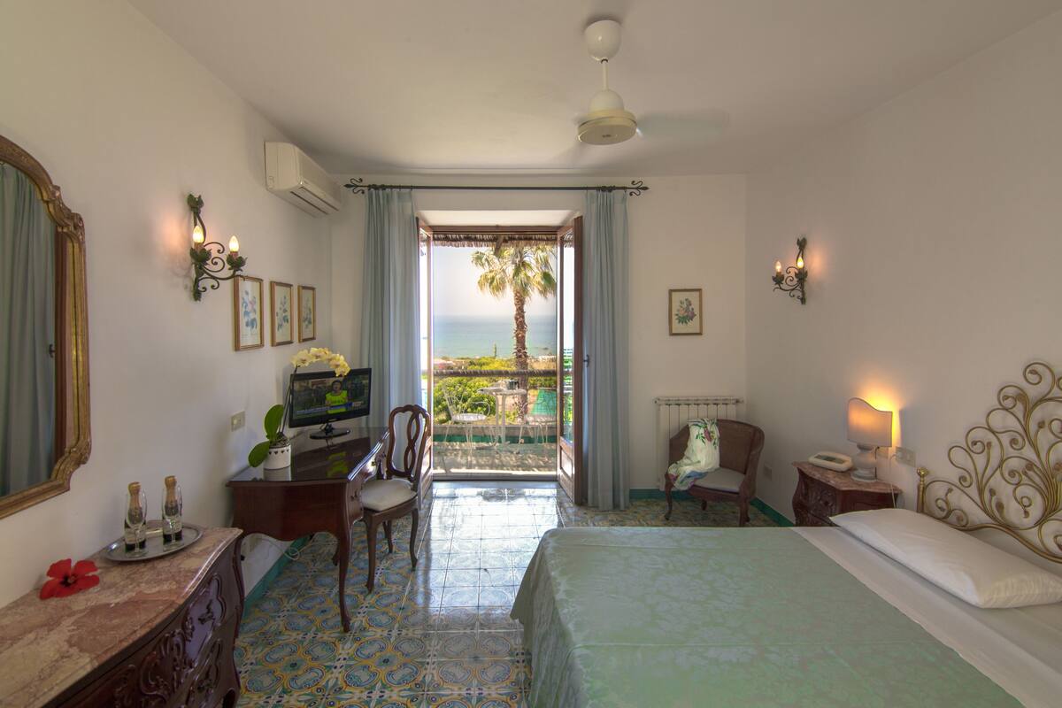 A hotel room with a bed, a desk with a computer, a chair, a small table, and a view of the ocean.