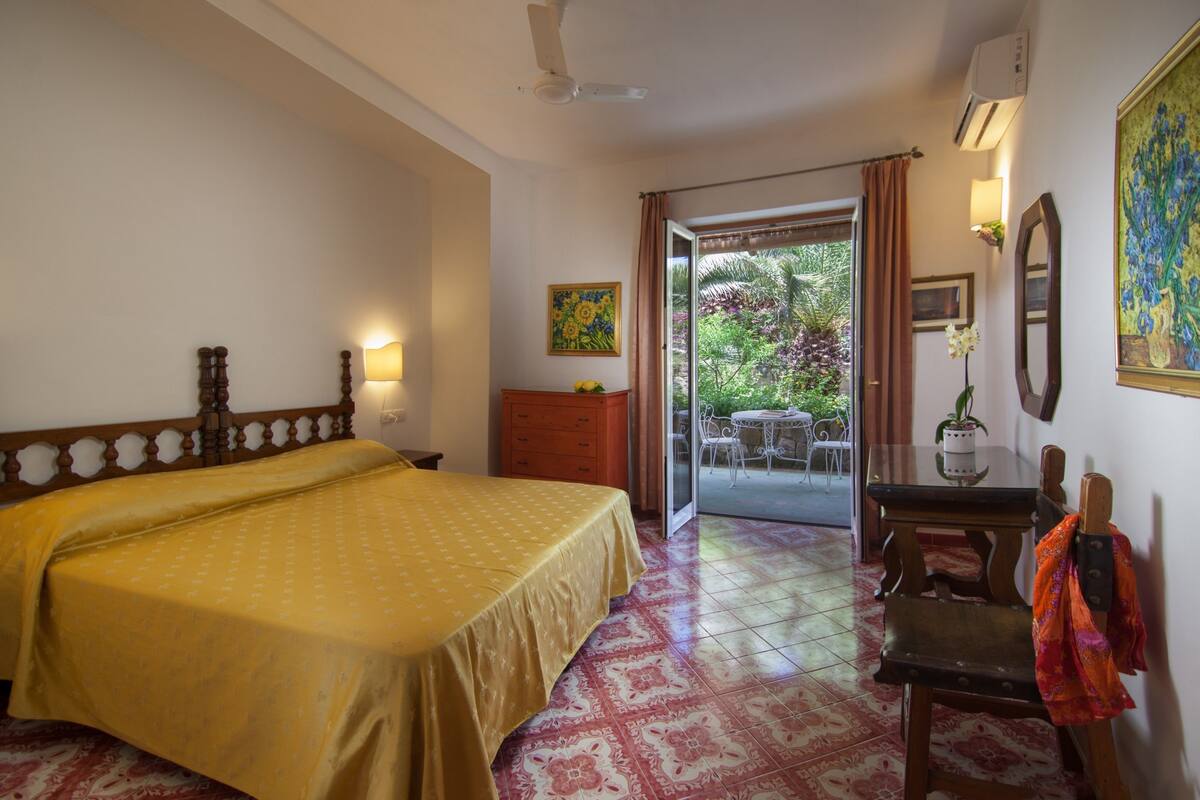 A hotel room with a bed, a desk with a chair, a dining table with chairs, and a view of the garden through the door.