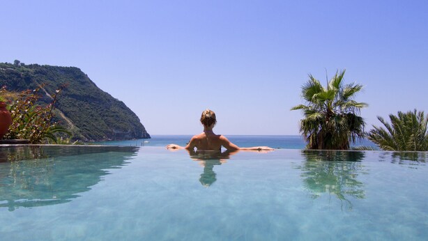A person relaxing in a clear infinity pool with a view of the ocean and a hillside. - Semiramis Hotel De Charme (Forio)