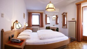 Habitación de hotel con una cama grande, cabecera de madera, lámparas de noche, una mesa con una silla, una ventana con cortinas, un radiador y suelo de madera.