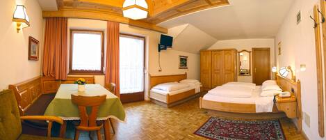 A room with a wooden bed, a small table with a green tablecloth, a chair, a wooden wardrobe, and a large window with curtains.