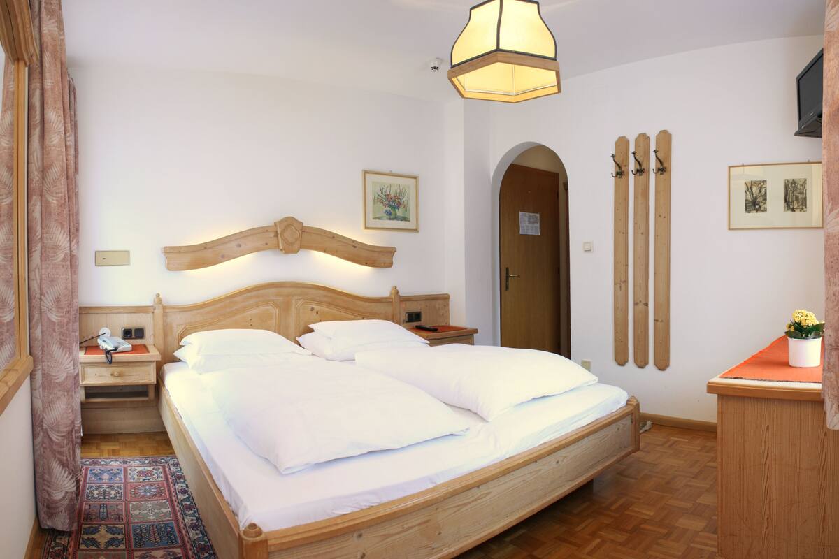 Un dormitorio con una cama de madera, una mesita de noche, un aparador, un ventanal con cortinas y una puerta de madera.