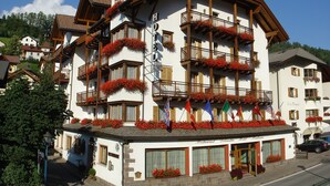Un hotel de varios pisos con balcones decorados con flores rojas, un letrero que indica que es restaurante, hotel y restaurante, y banderas que representan diferentes países.