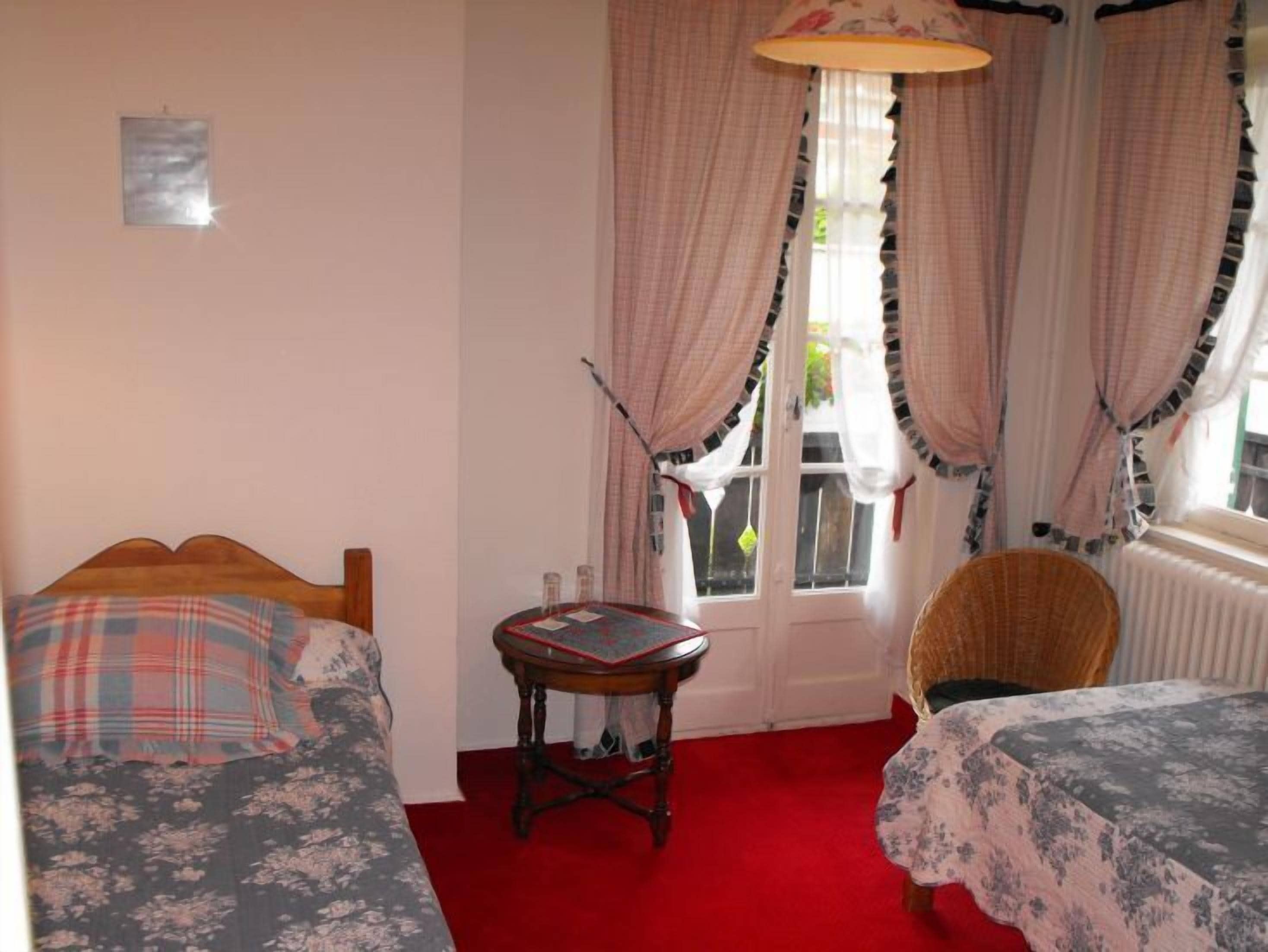 A room with two beds, a small table, and a chair. There is a window with curtains and a radiator.