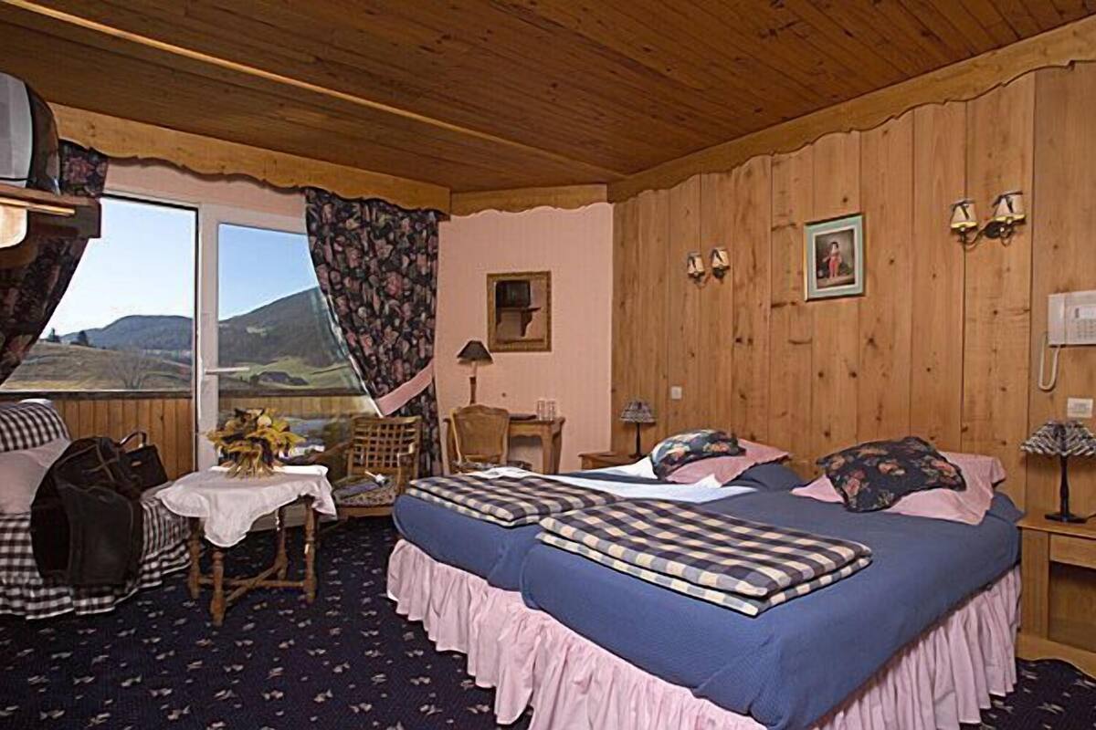 A cozy bedroom with two beds, a wooden ceiling, a window with curtains, a table with a vase, and a chair.
