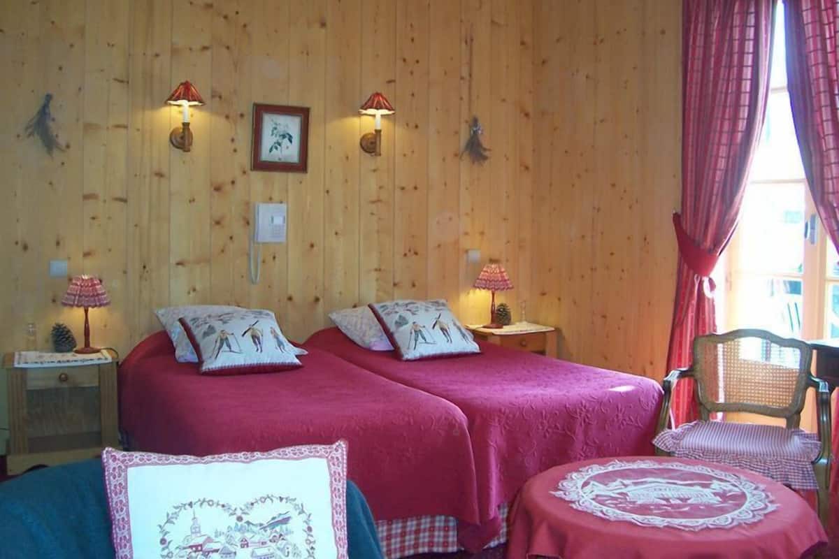 A room with two beds, wooden walls, and a window with red curtains.