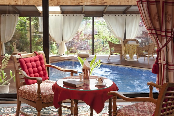 A cozy seating area with a pool view, featuring a wooden chair with a red cushion, a small round table with a red tablecloth, and a potted plant.